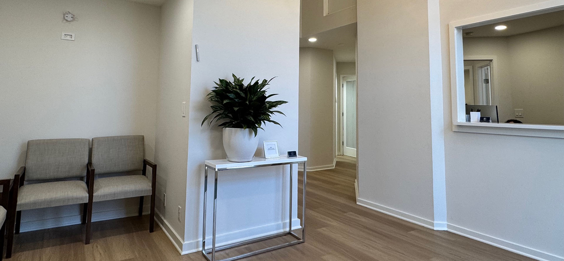 The image shows a well-lit room with a waiting area featuring seating, a reception desk, a potted plant, and a wall-mounted sign.
