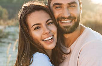 The image shows a man and a woman embracing each other outdoors during daylight with a clear sky, both smiling and appearing happy.