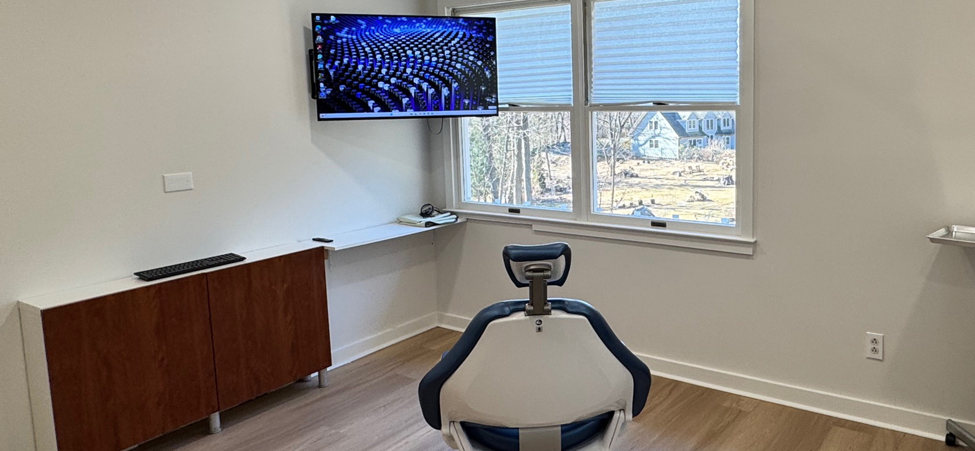 The image shows an interior space with a modern office setup featuring a desk with a computer monitor, a rolling chair on wheels, a window with blinds partially drawn, and a large screen displaying a colorful graphic.