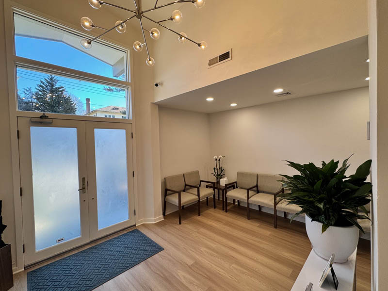The image shows a modern interior space with a waiting area featuring a wooden floor, a white rug, a beige sofa, a wooden bench, a coffee table, a potted plant, a large window with frosted glass, and a ceiling light fixture.