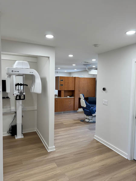The image shows an interior view of a dental office waiting area, featuring a modern dental chair with a digital display, a ceiling-mounted light fixture, a sleek reception desk, and a clean, minimalist aesthetic with a warm color palette.