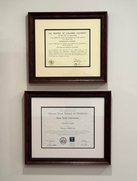 The image shows two framed certificates hanging on a wall, each with text indicating an educational achievement, specifically a diploma from Columbia University and a degree from New York University.