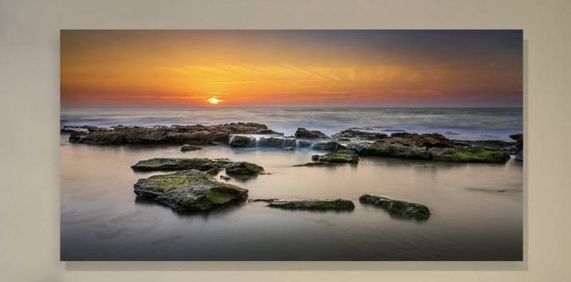 The image is a photograph depicting a serene coastal scene at sunset with calm waters reflecting the sky, and rocks jutting out of the water near a beach.