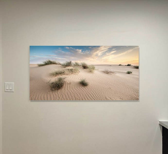 The image shows a large canvas print of a desert landscape with sand dunes at sunset, displayed on a wall.
