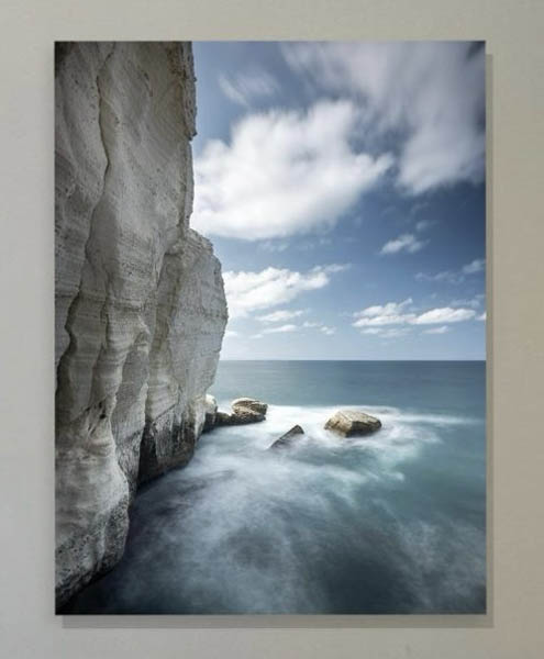 The image depicts a rugged coastal landscape with large, white rock formations rising from the ocean, featuring clear blue skies and calm waters.