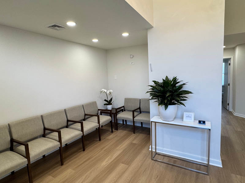 The image shows a waiting area inside a medical office with a modern design, featuring beige walls, a white ceiling, a clean floor, a potted plant on a small table, several chairs arranged around a reception desk, and a bench.