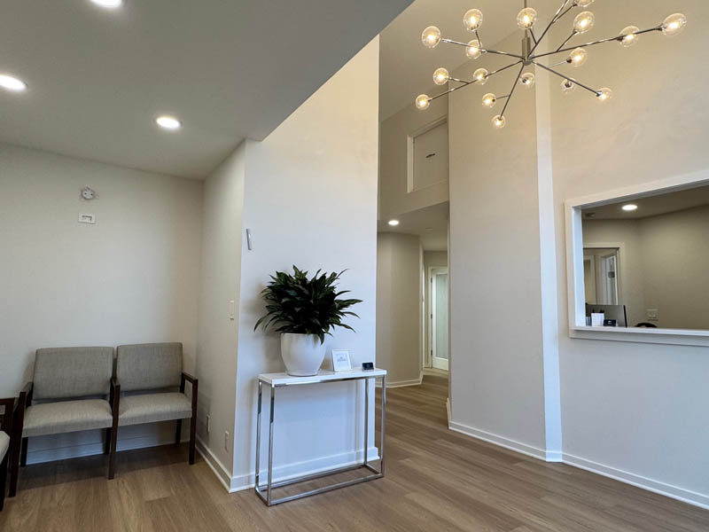 The image shows an interior space with a modern design, featuring a waiting area with a bench, a reception desk with a plant on top, a chandelier hanging from the ceiling, and a large window allowing natural light into the room.
