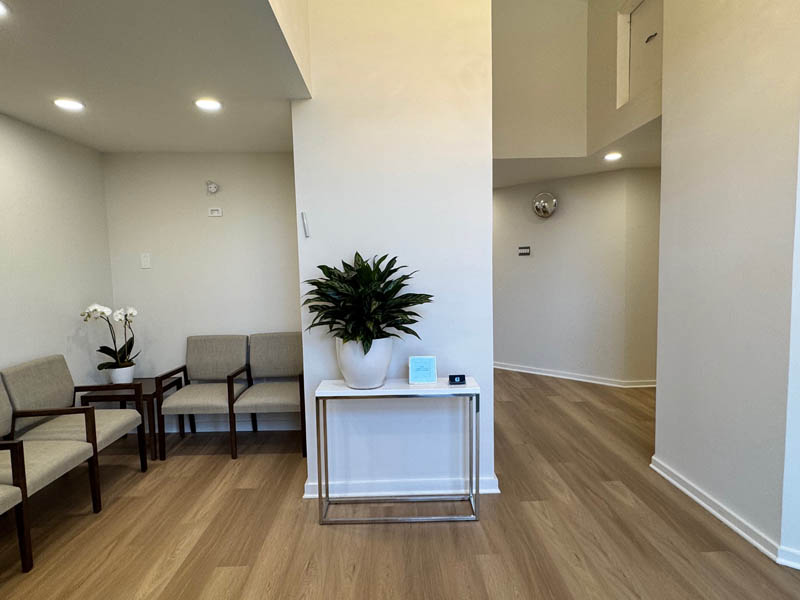The image shows an interior space with a modern design, featuring a waiting area within a medical facility. There are white walls, a light-colored hardwood floor, and a minimalist decorative style. A potted plant sits on a small table, and there s a reception desk in the corner. The room is well-lit and appears clean and organized.