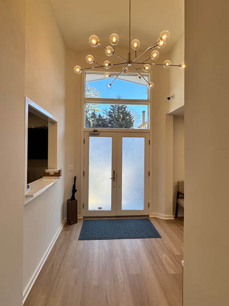 The image shows an interior view of a room with a modern design, featuring a large glass door at the end of a hallway, a light fixture hanging from the ceiling, and a white wall with a decorative light fixture on it.