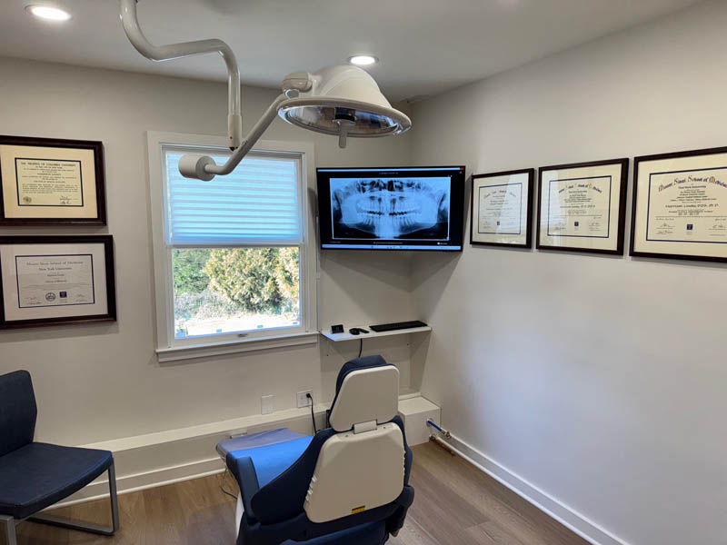 The image shows an interior view of a dental office with a dental chair, examination equipment, and framed certificates on the wall behind the chair.