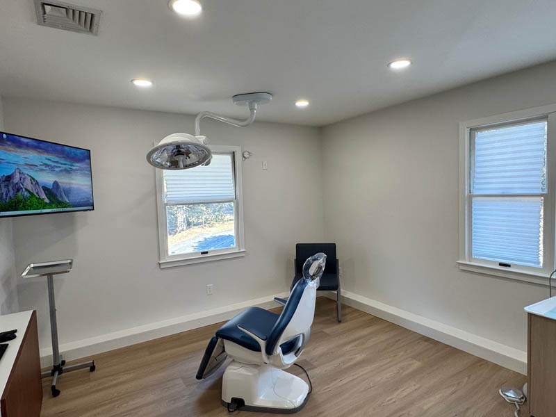 The image shows a modern dental office interior with a treatment chair, a monitor displaying a natural landscape, and a window with blinds partially drawn.